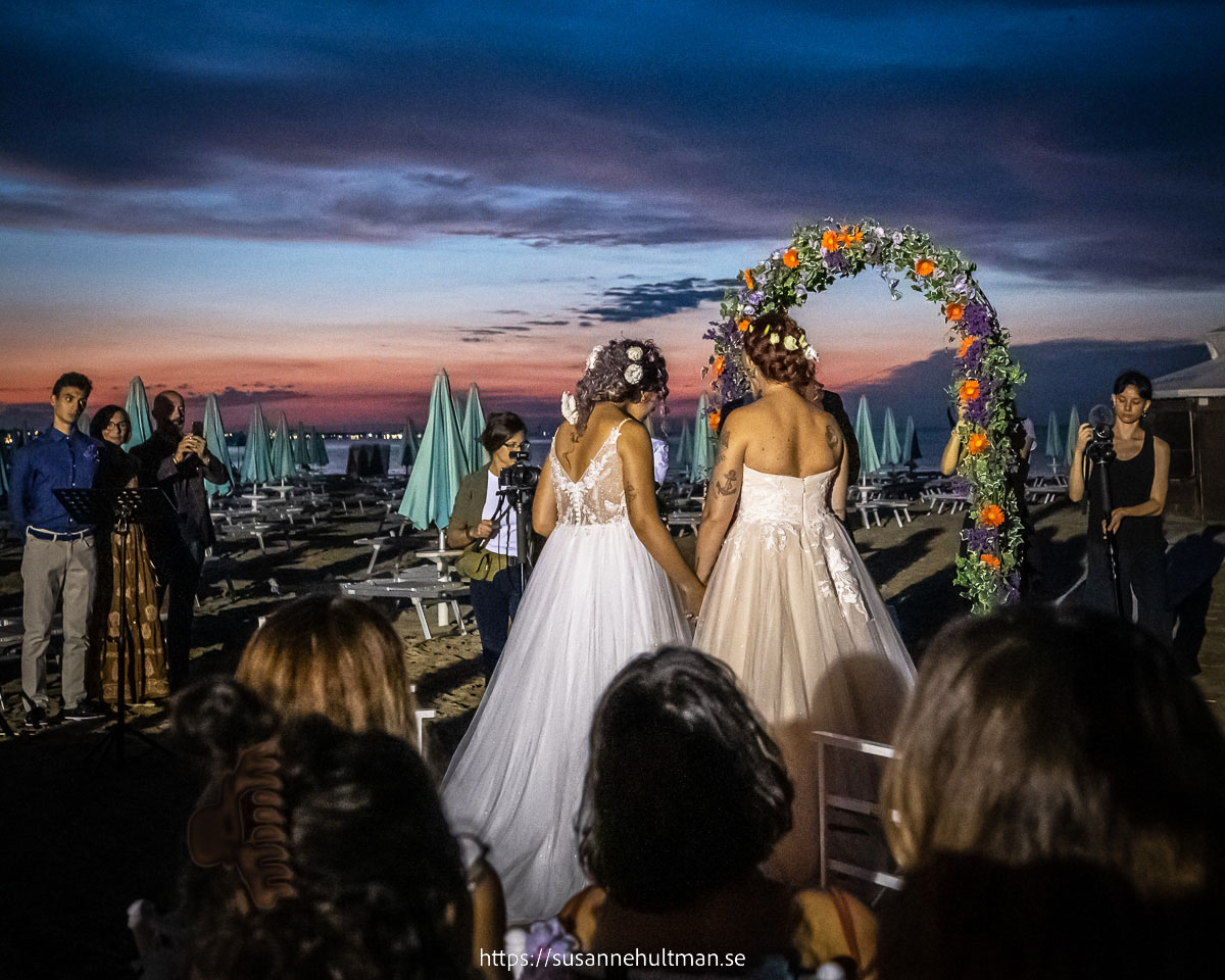 Två kvinnor i brudklänningar med ryggen mot kameran framför en blomsterbåge och solnedgångshimmel.