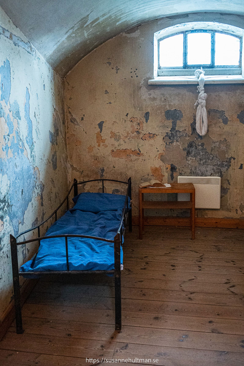 Fängelsecell med säng och sängbord nedanför fönster.