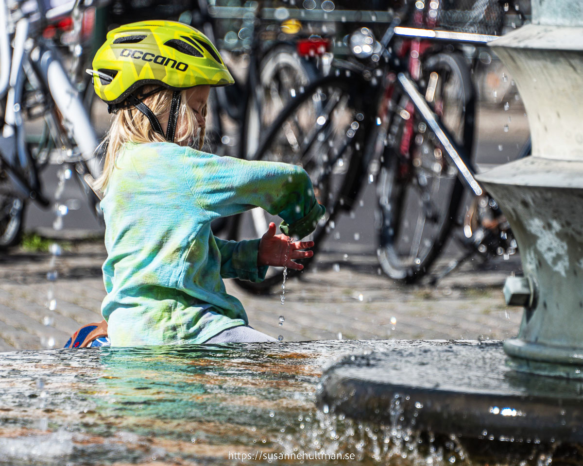 Flicka i gul hjälm som leker med vattnet i en fontän.
