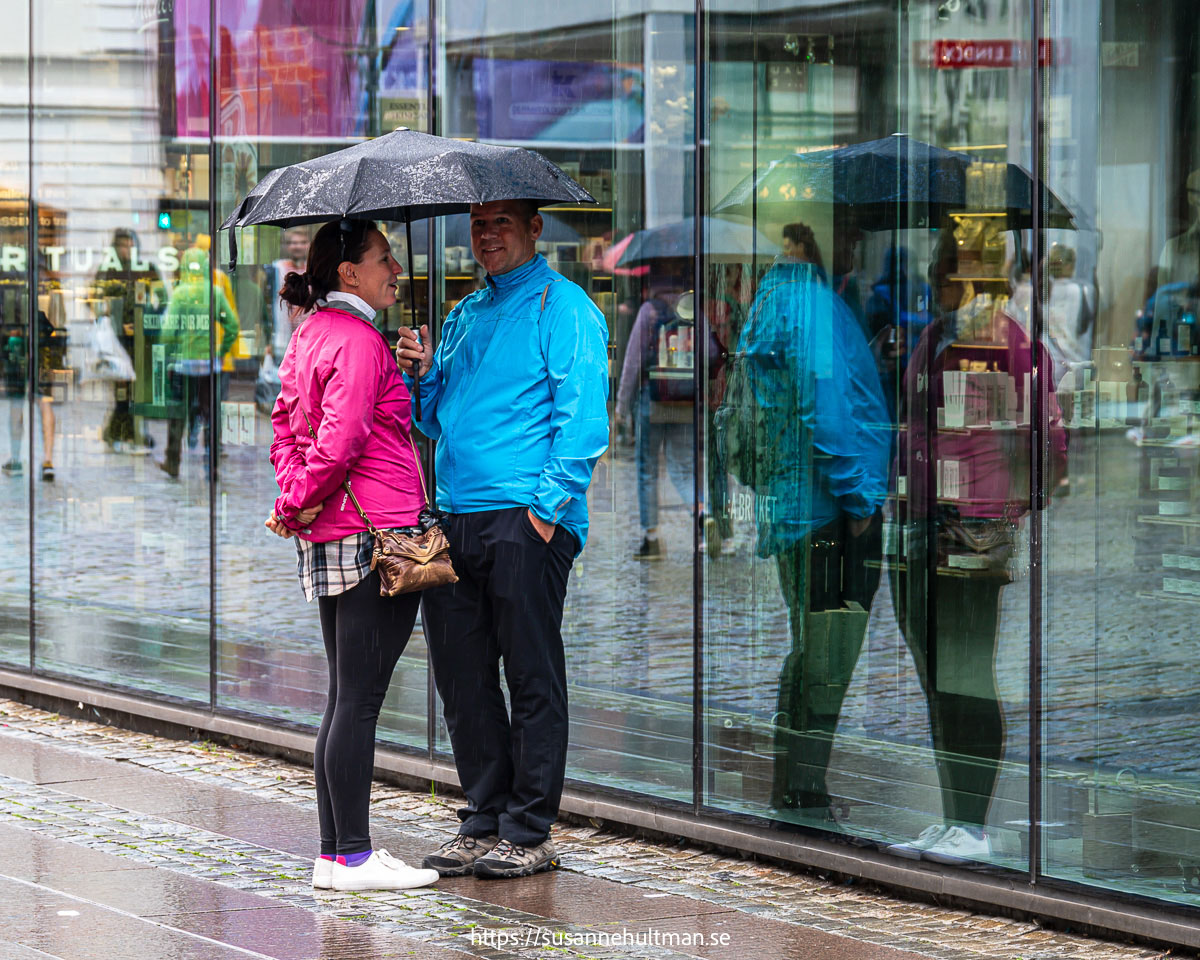 Ett par står under ett paraply och deras spegelbild syns i ett skyltfönster.