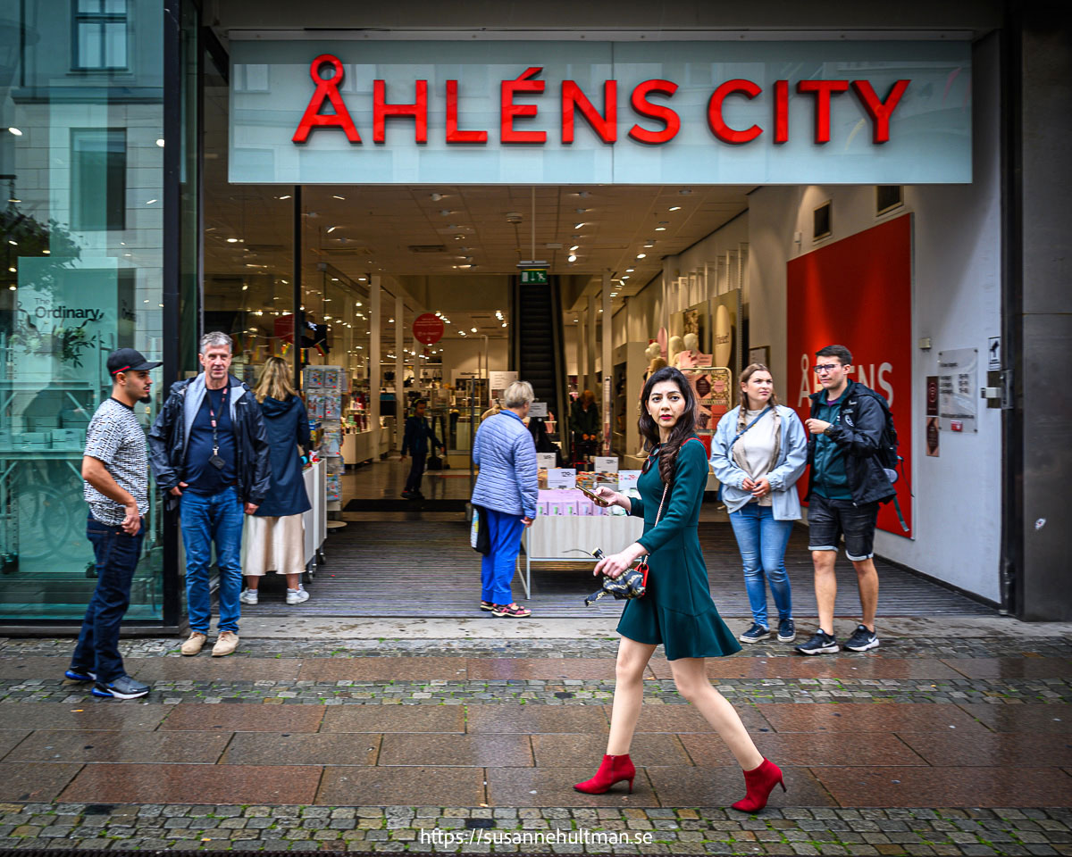 Kvinna i grön klänning och röda skor går förbi ingången till Åhléns där det står många andra personer.