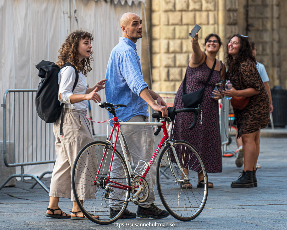 Man leder cykel och kvinna bredvid. I bakgrunden två kvinnor med mobil lyft.
