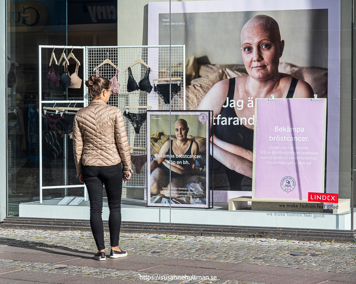 Kvinna vid skyltfönster med reklam för bh:ar och kvinna med kalt huvud och skylt med "Bekämpa bröstcancer".