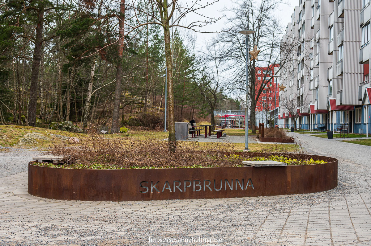 Texten "Skarpbrunna" på låg kant runt plantering med ett träd.