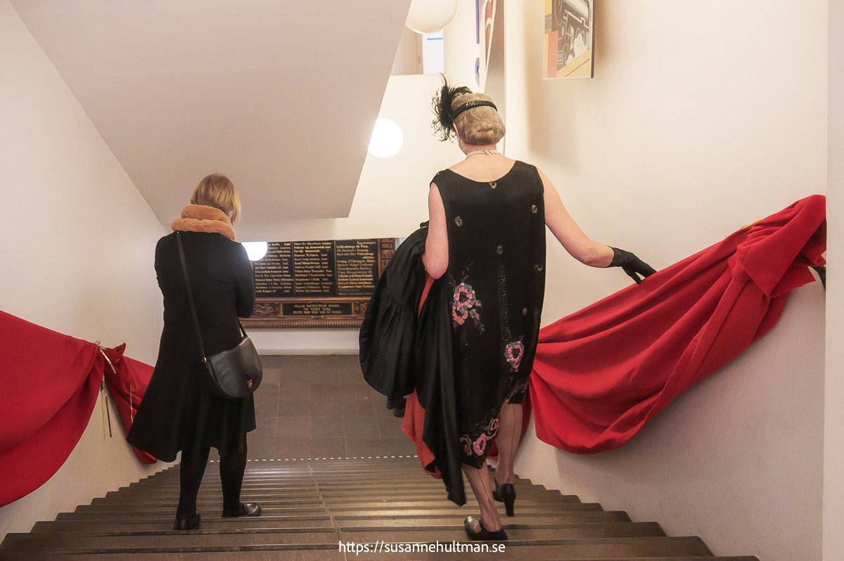 Två kvinnor går nerför en bred trappa. En av dem i kläder från 1920-talet. Röda tyg draperar trappräckena.