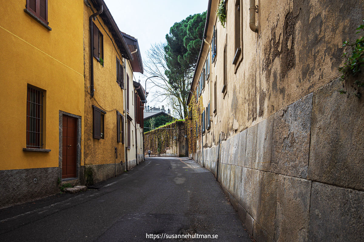 Vägen till Malpensata mellan gamla byggnader. Till vänster en gulfärgad och till höger en brungrå med hög stensockel.