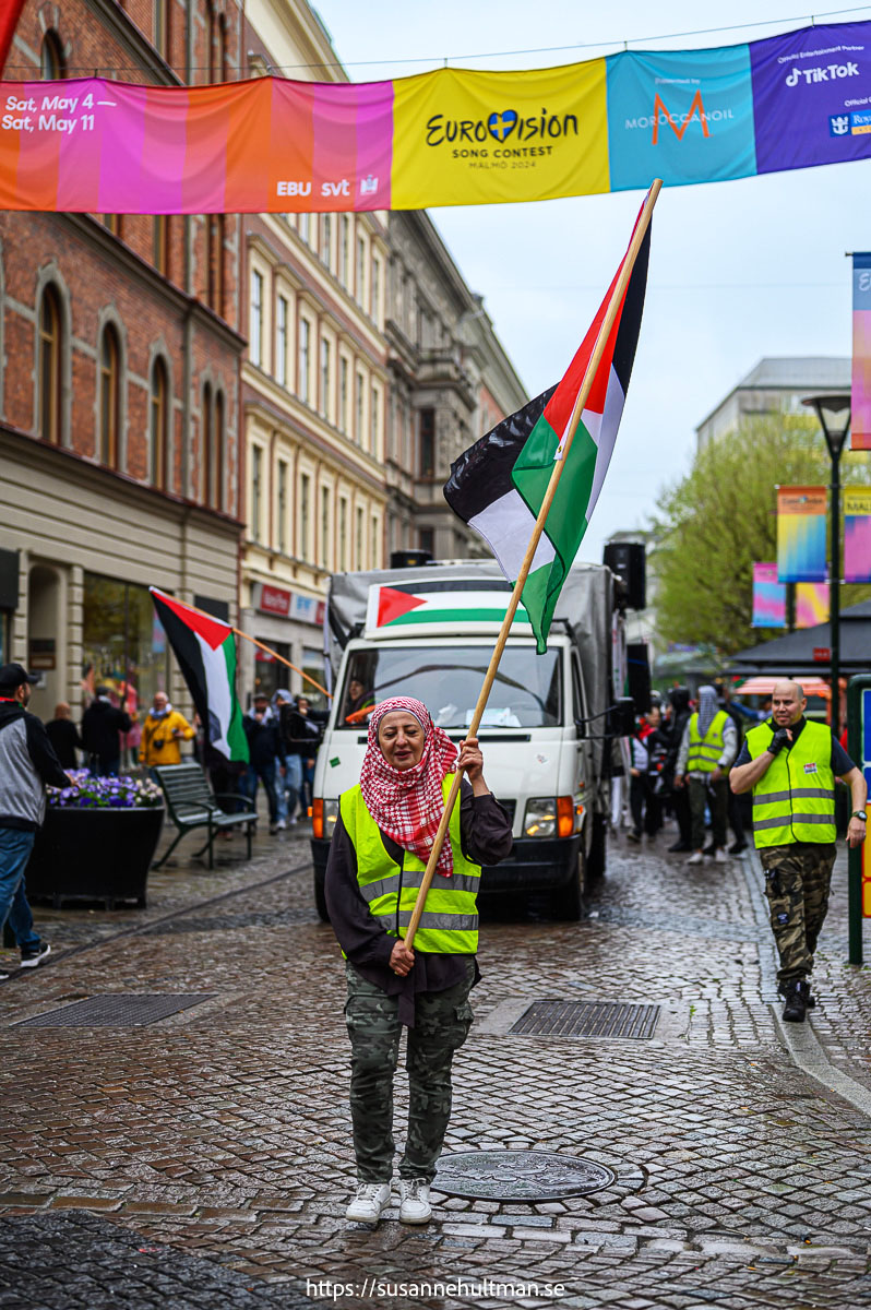 Kvinna med palestinaschal och palestinaflagga under banderoll med reklam för Eurovision.